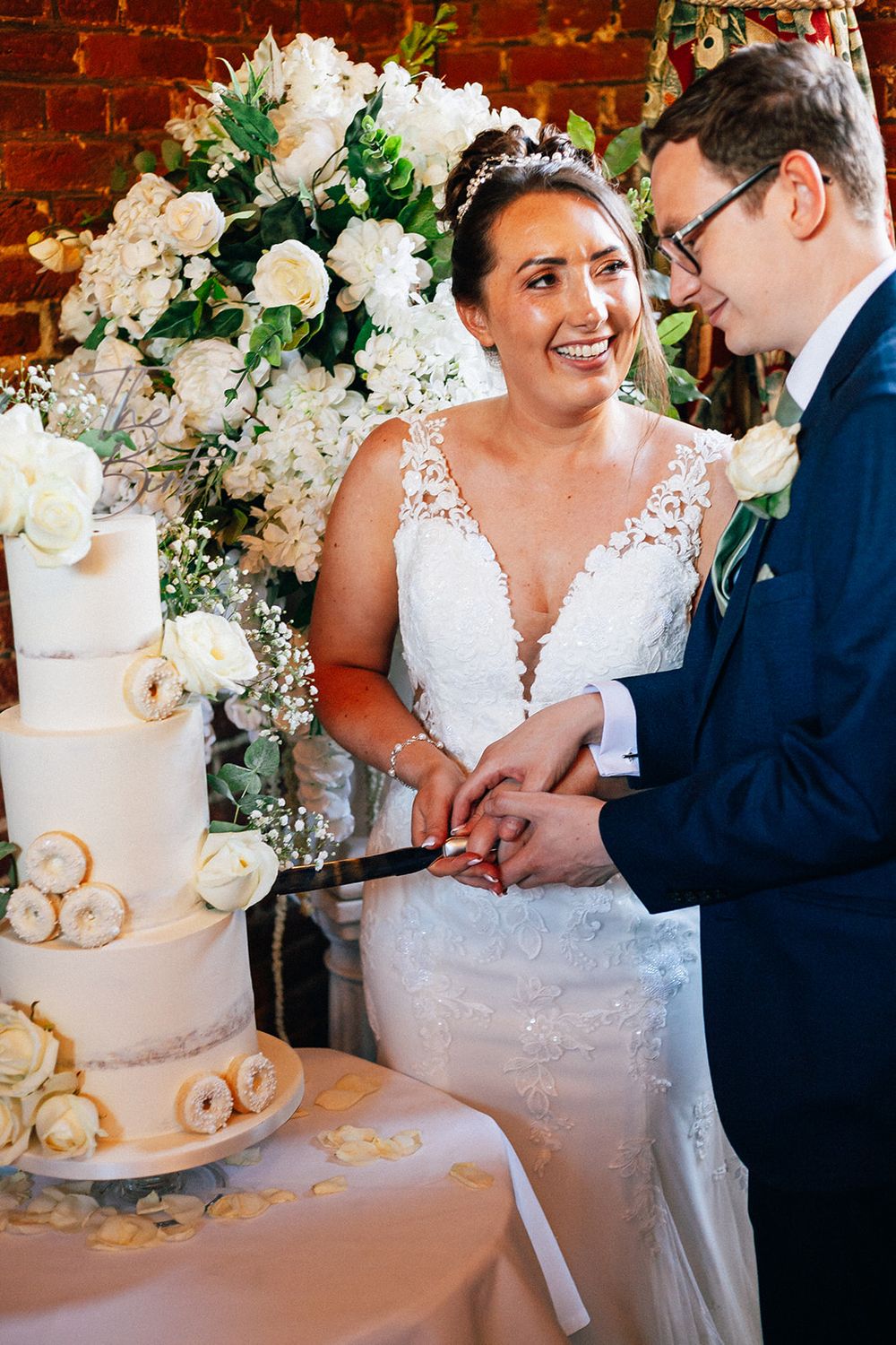 A couple cutting their elegant white wedding cake decorated with fresh roses and greenery at a rustic brick venue.