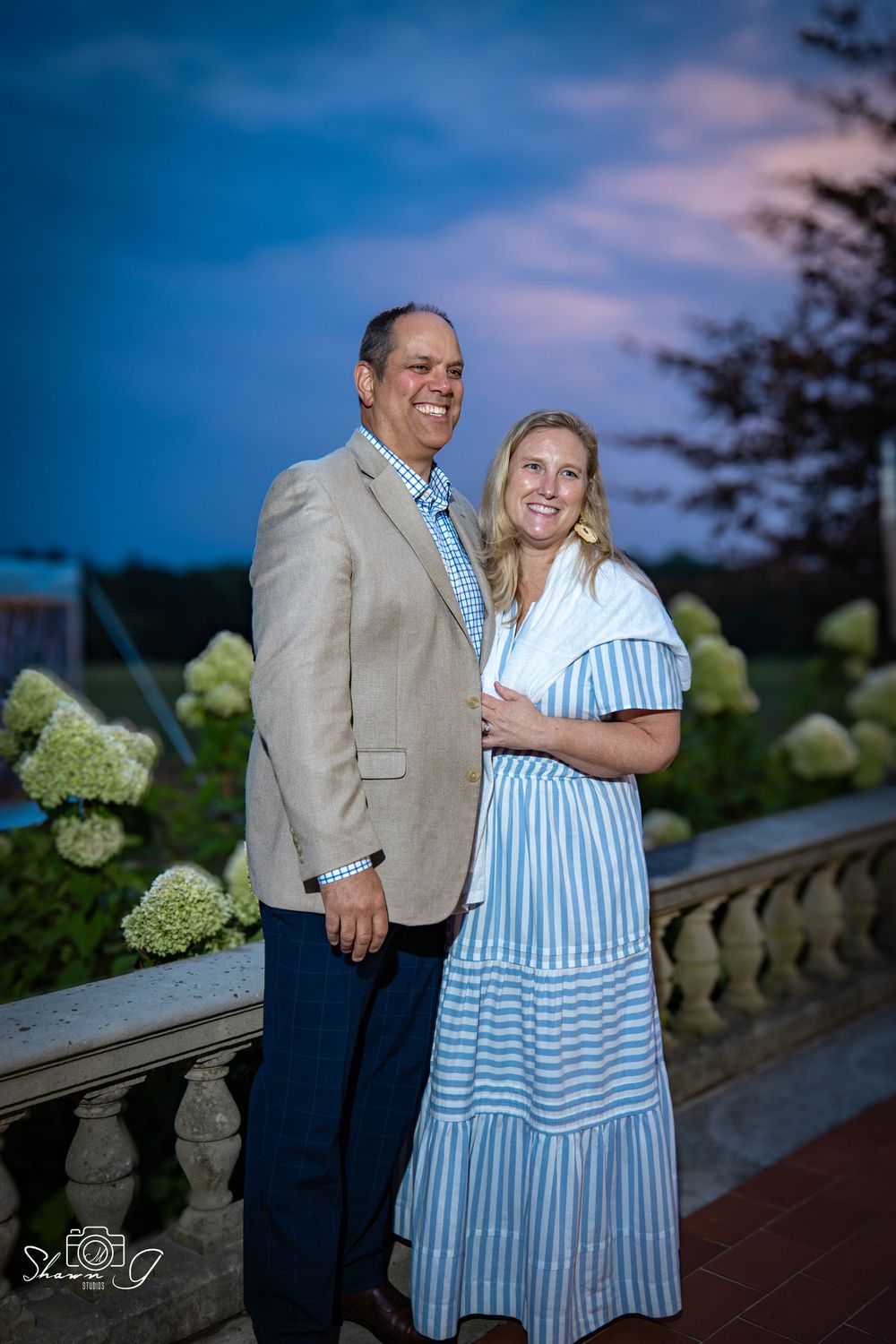 A couple poses together on a terrace at dusk, with hydrangea bushes and a blue sky in the background.