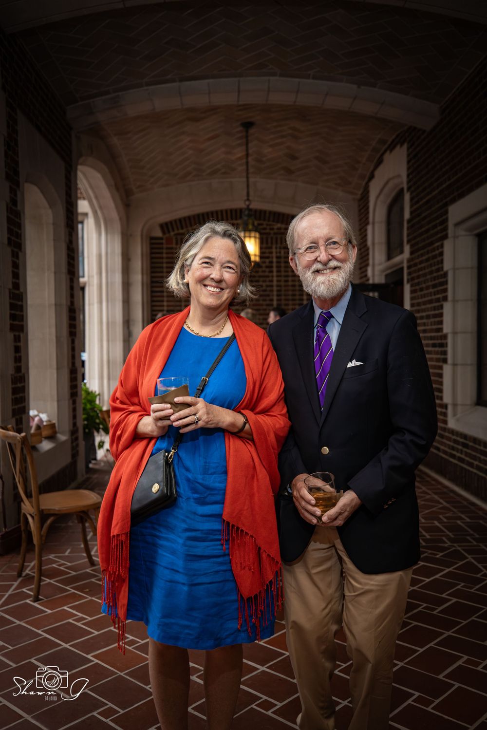 A couple in formal attire stands in an arched brick hallway with warm lighting and patterned tile floors.