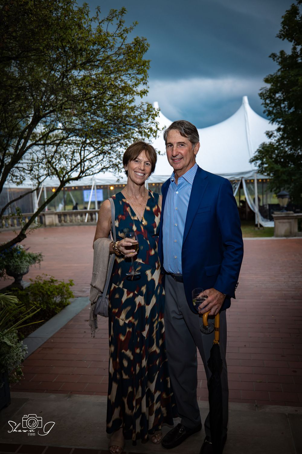 A couple stands together on a wooden deck during a cloudy day, with the woman wearing a patterned dress and man in blue suit.