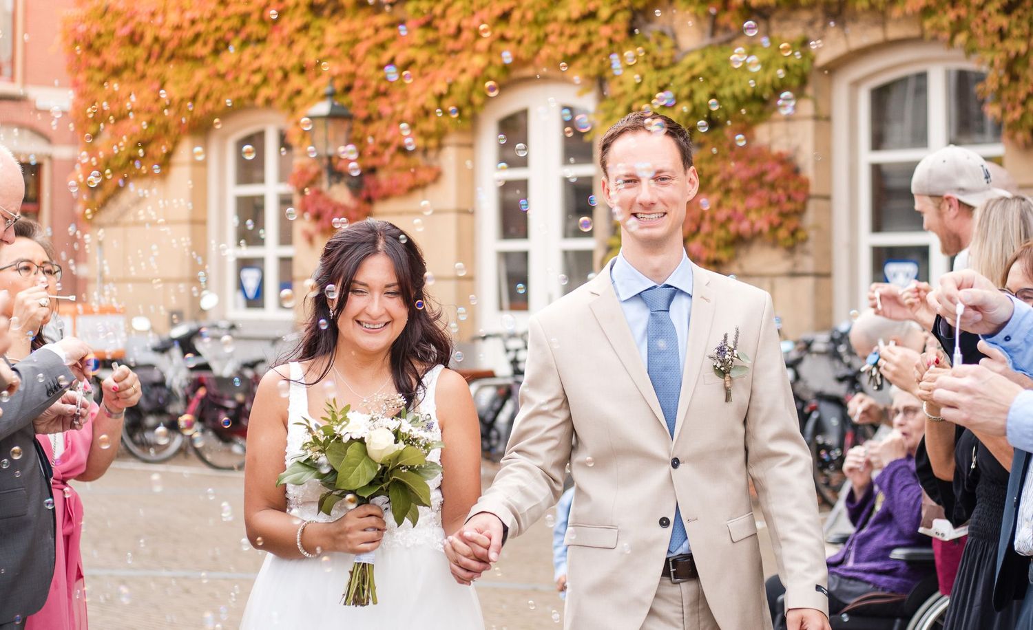 A wedding party celebrates with soap bubbles and laughter outside a historic brick building on an autumn day.