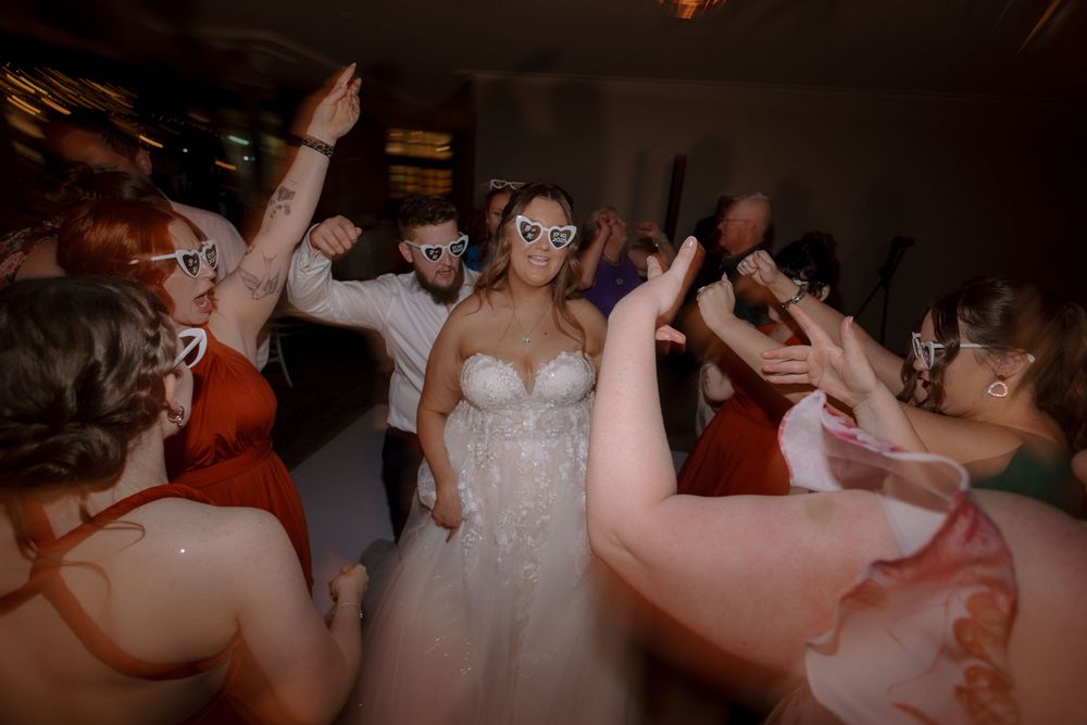 A group of people dancing and celebrating at a wedding reception on a dark dance floor.