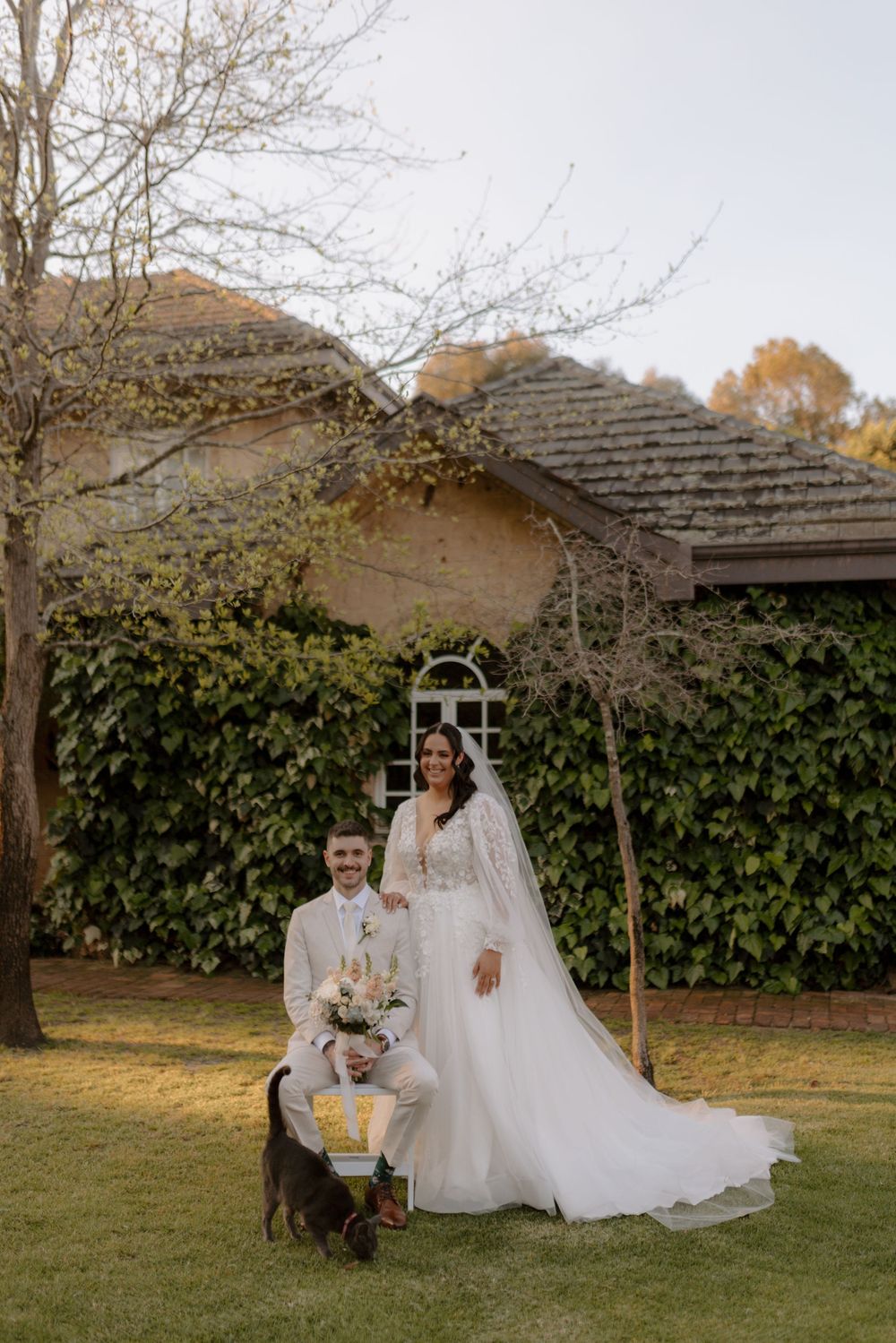 Outdoor wedding scene with couple standing under rustic arch with ivy-covered wall and autumn trees in background.