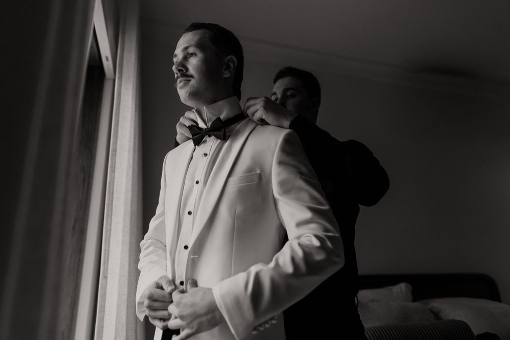 A black and white photograph showing a groom in a white tuxedo jacket standing by a window.