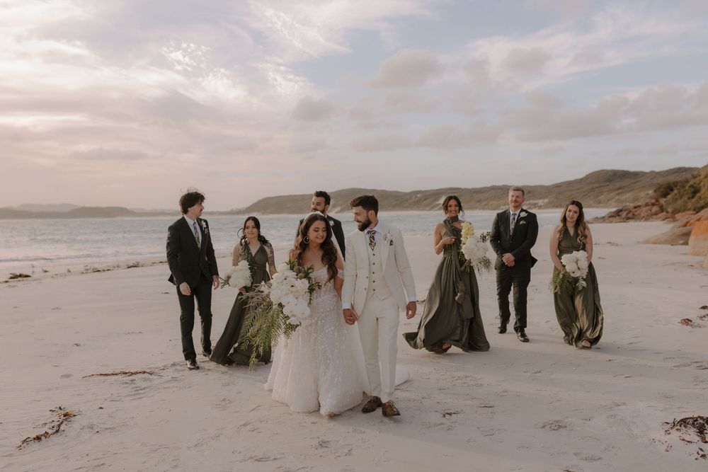 Wedding party walking together along a scenic beach with sand dunes and ocean views at sunset.