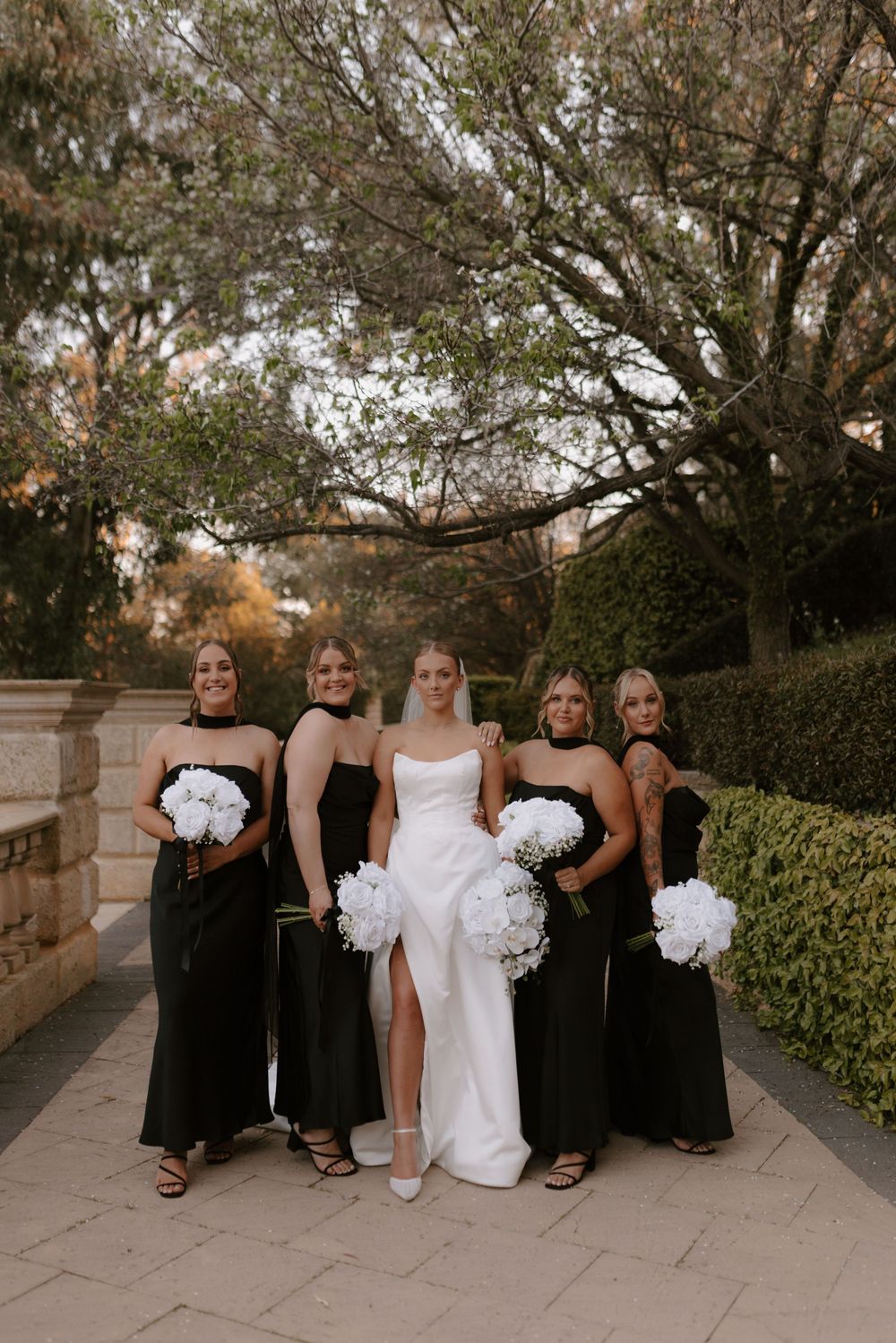 Bridal party in black dresses and white gown sitting on stone steps during outdoor wedding photo session.