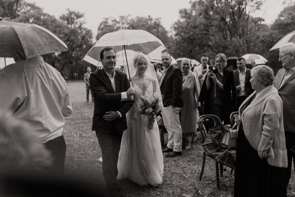 Wedding guests huddle under umbrellas during an outdoor ceremony on a rainy day.