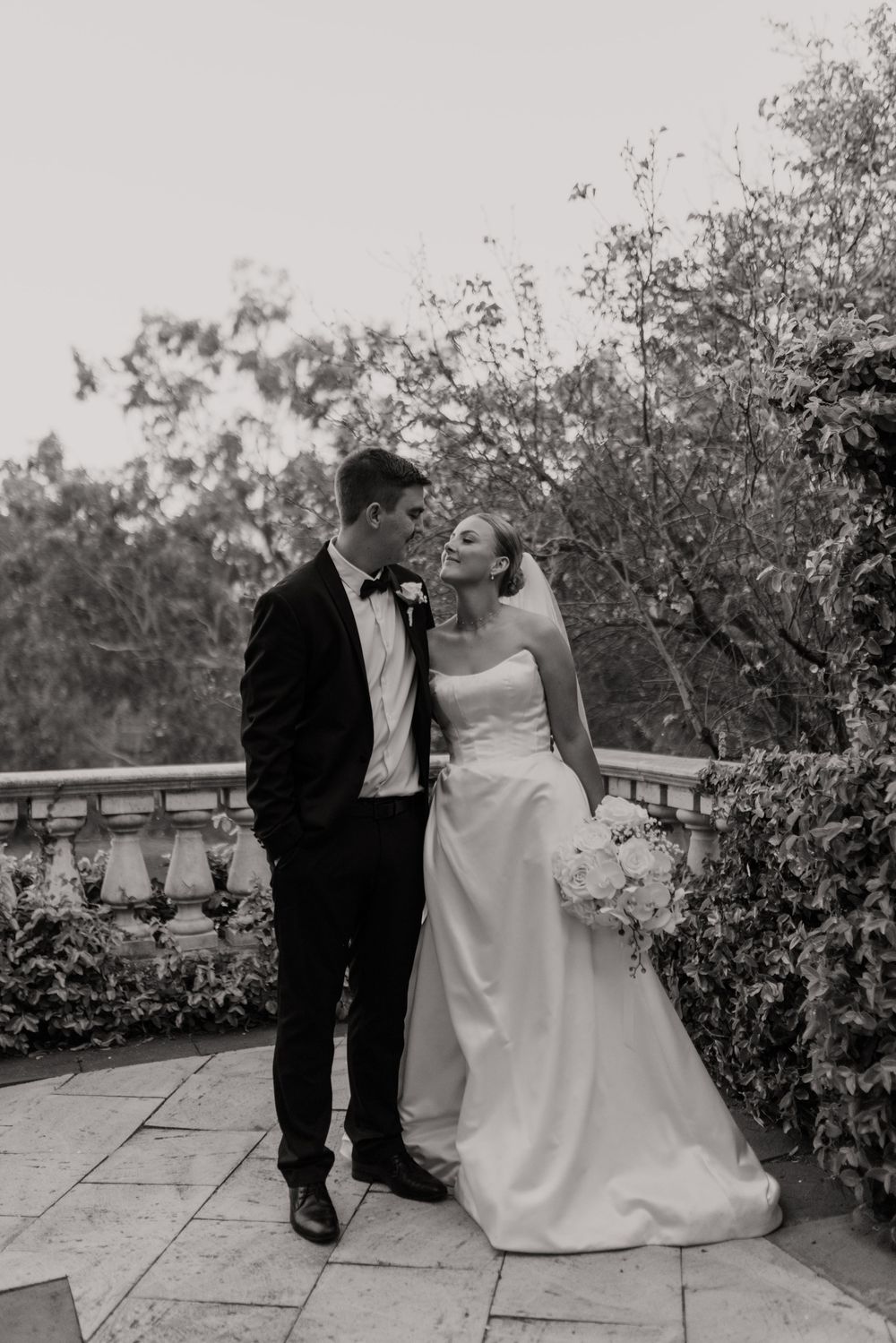 A romantic black and white wedding photo series showing a couple seated on stone steps surrounded by garden foliage.