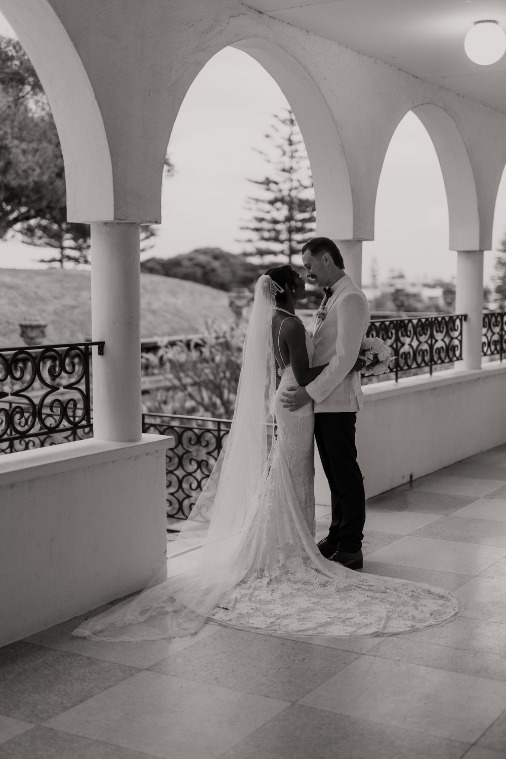 A romantic wedding moment captured under elegant white arches with ornate railings and a flowing cathedral train dress.