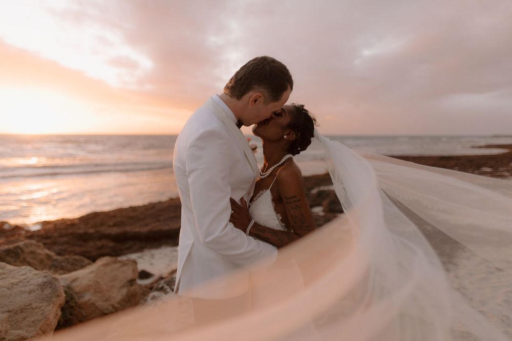 Newlywed couple embraces on a beach at sunset with flowing white dress and dramatic coastal backdrop.