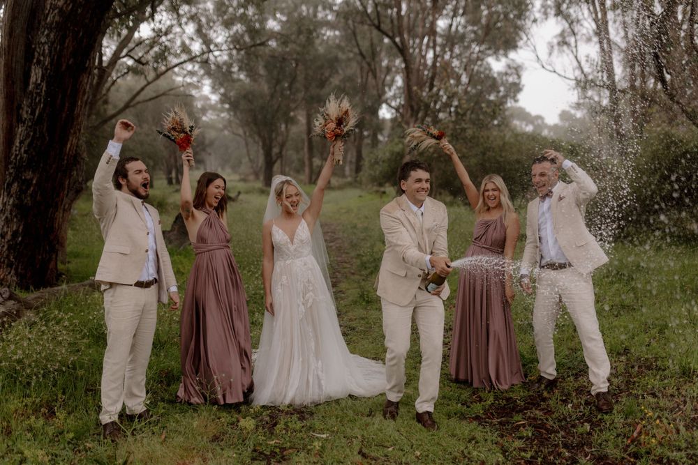 A group in wedding attire poses together in a natural outdoor setting with trees and greenery in the background.