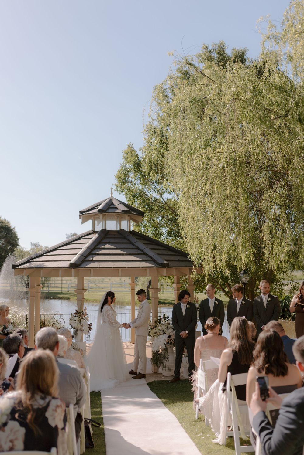An outdoor wedding ceremony takes place under a gazebo surrounded by willow trees on a sunny day.