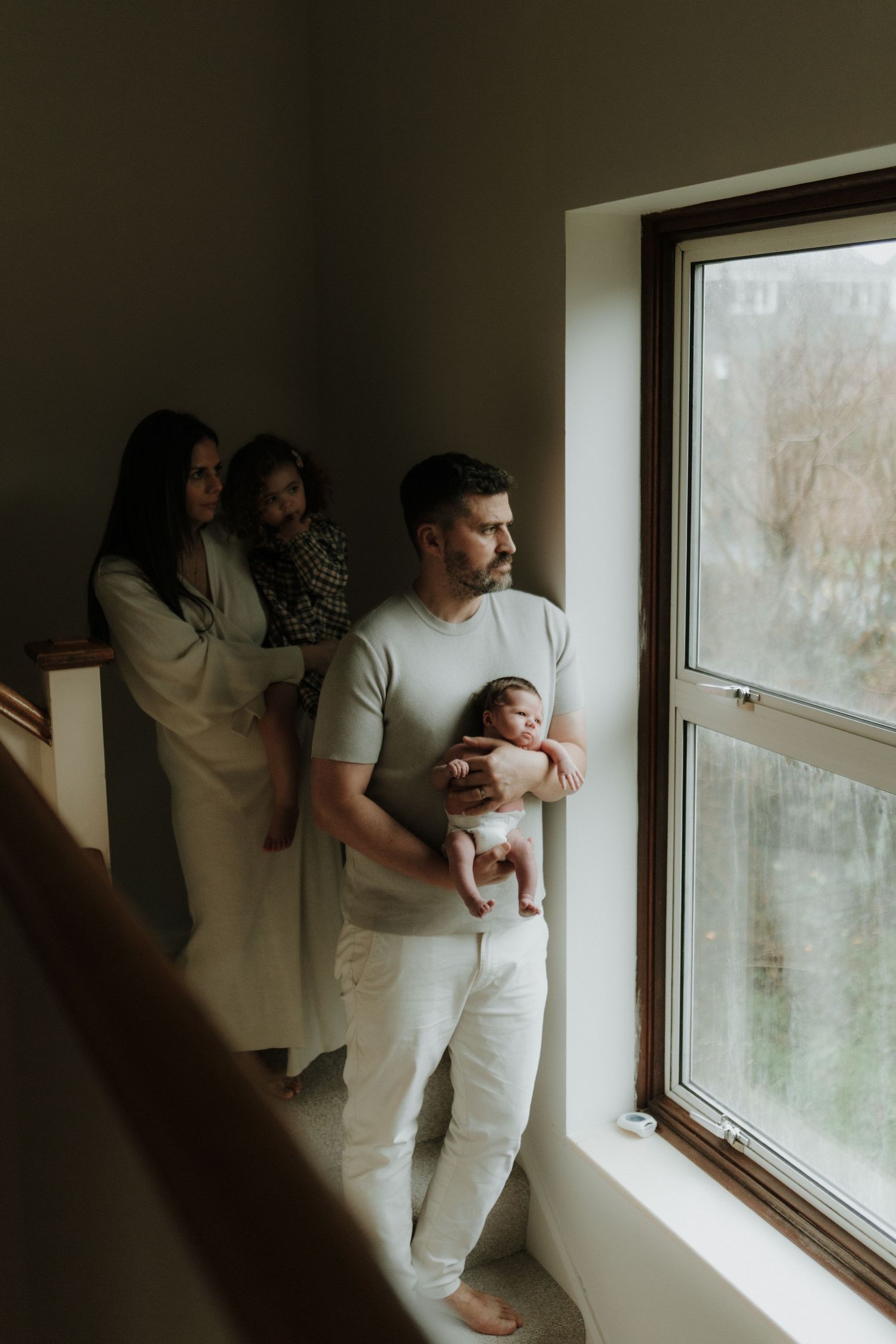 A family stands by a window in moody lighting, with a parent holding a newborn baby while others look on from behind.