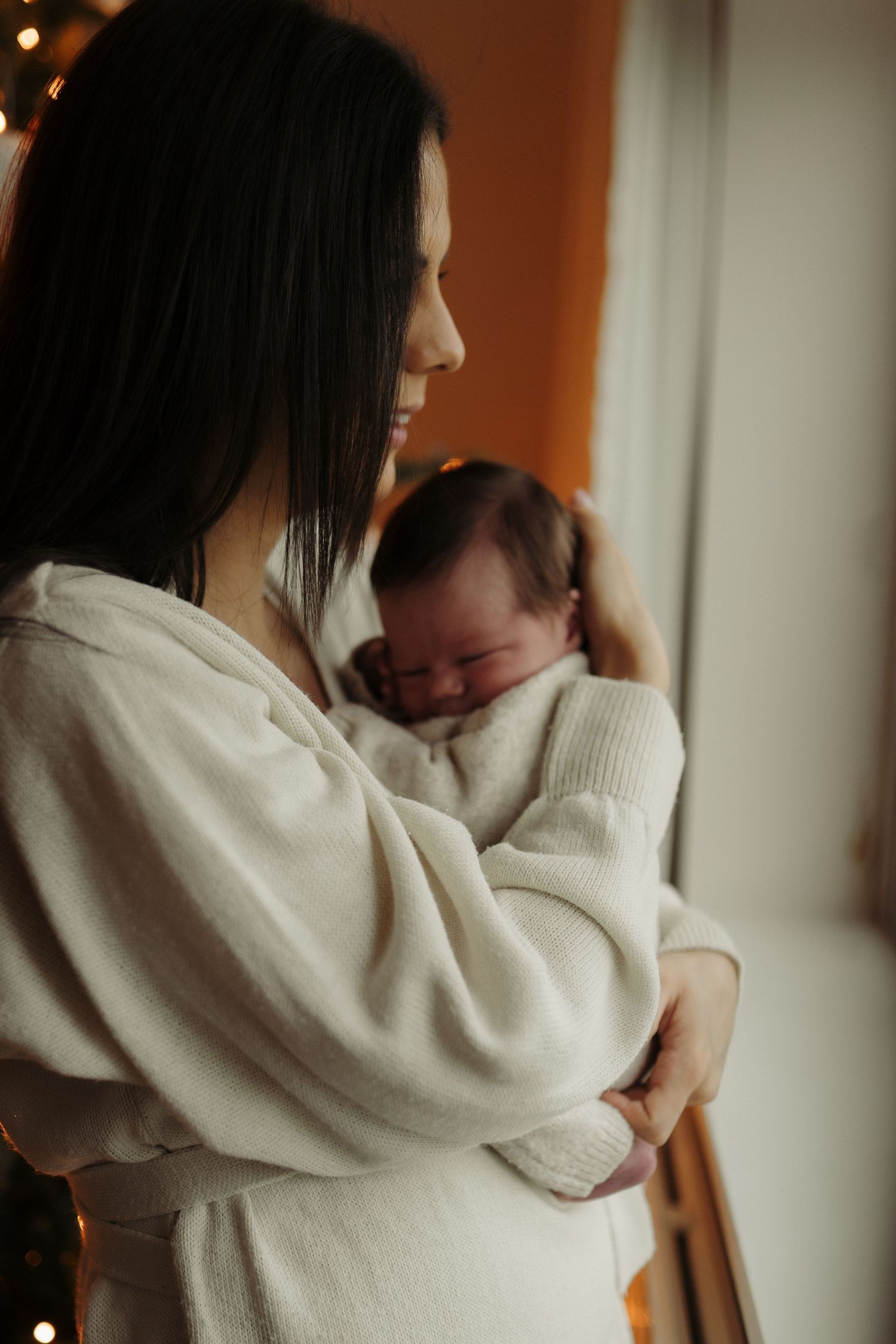 A person in a white sweater holds and comforts a small baby in a tender embrace by a window.
