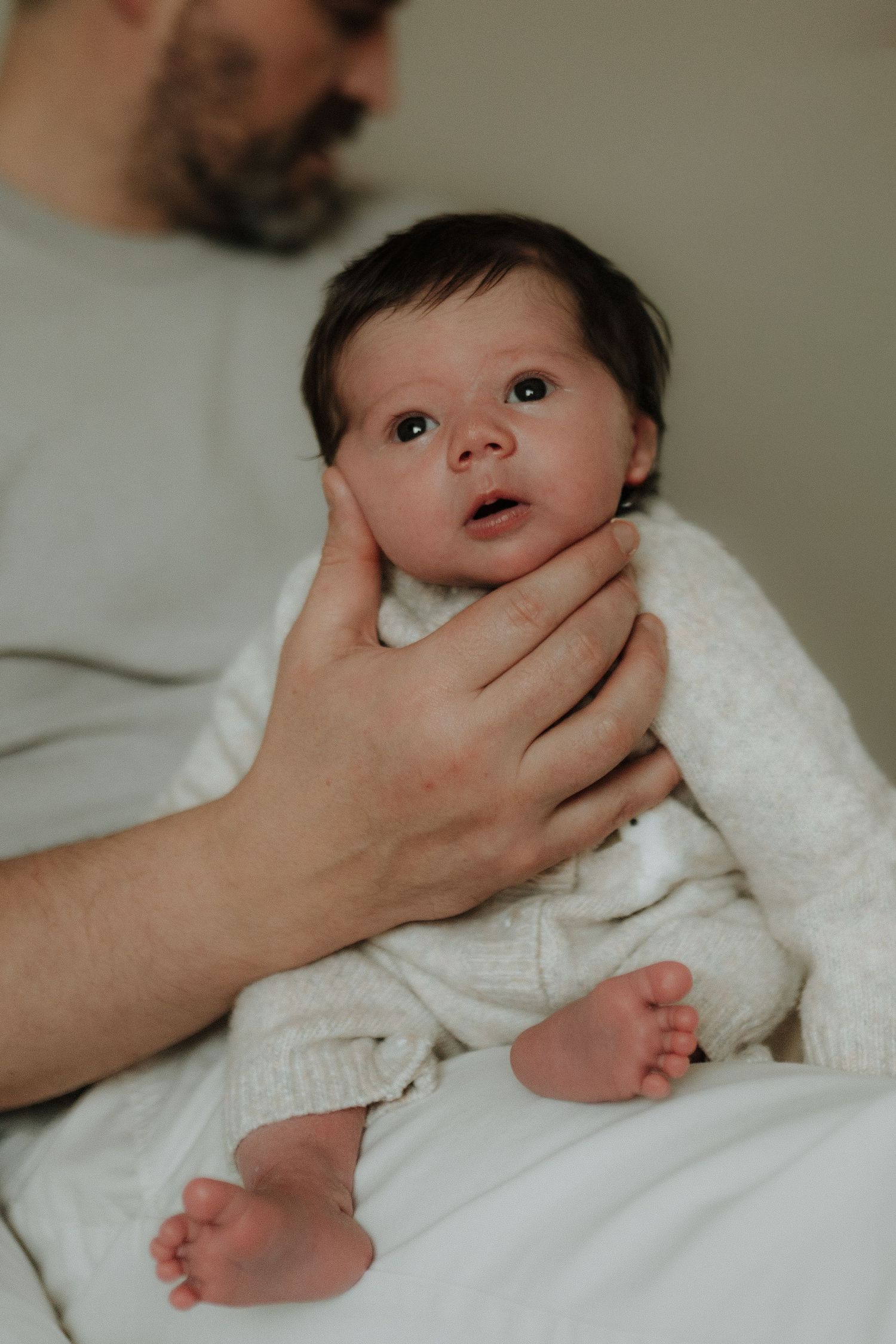 A gentle hand cradles a baby wearing a white sweater while lying on soft bedding.