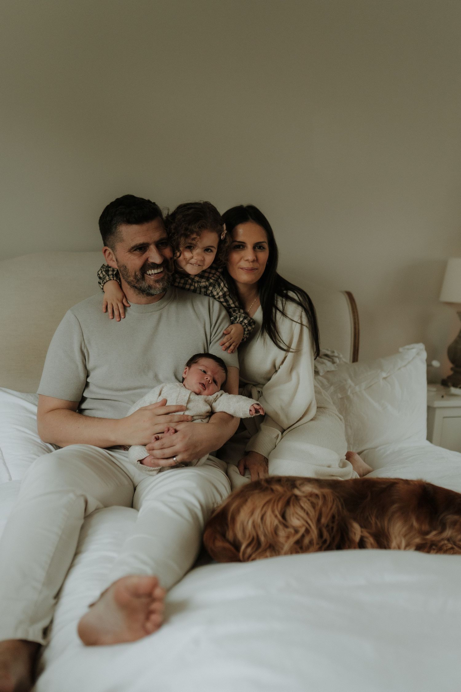 A family lounges together on a cozy white bed in matching neutral-toned outfits during an intimate lifestyle photoshoot.