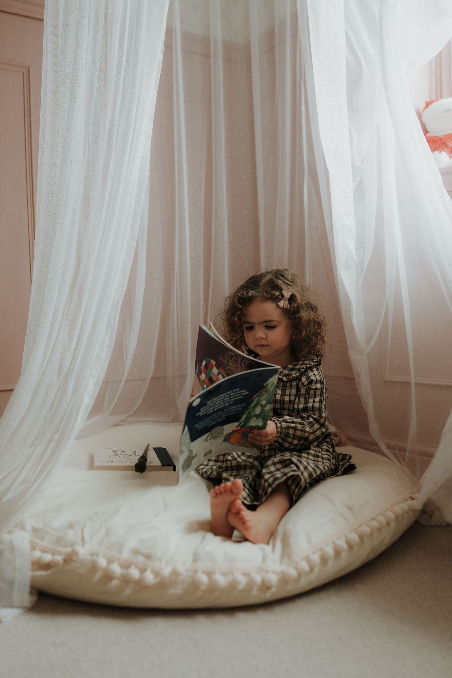 A child rests on a cozy floor cushion beside flowing white curtains in a minimalist room setting.