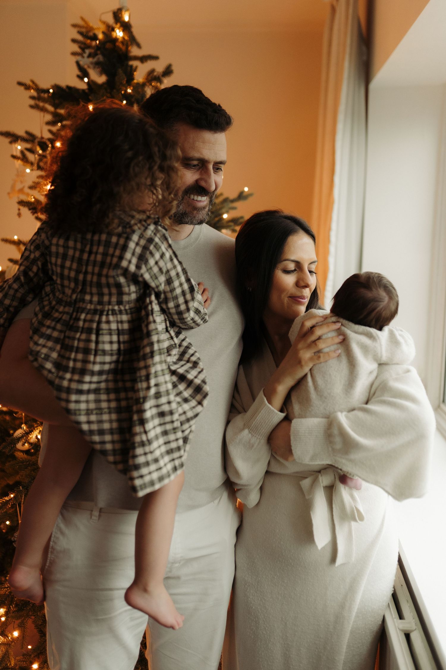 A family shares tender moments and laughter in front of a Christmas tree in a warm-toned series of candid photos.