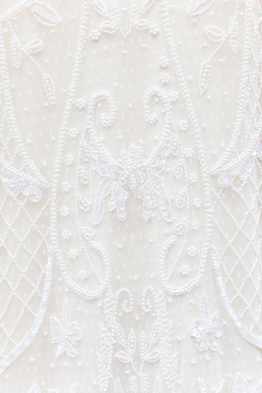 Intricate white beaded lace pattern with floral and geometric designs on wedding dress fabric.