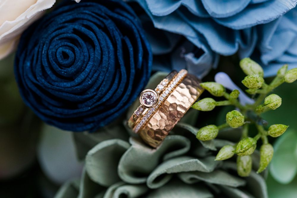 Wedding rings nestled among navy blue and light blue fabric flowers with green foliage.
