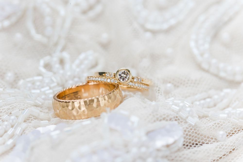 Gold wedding rings with intricate details photographed on white lace fabric in soft natural lighting.