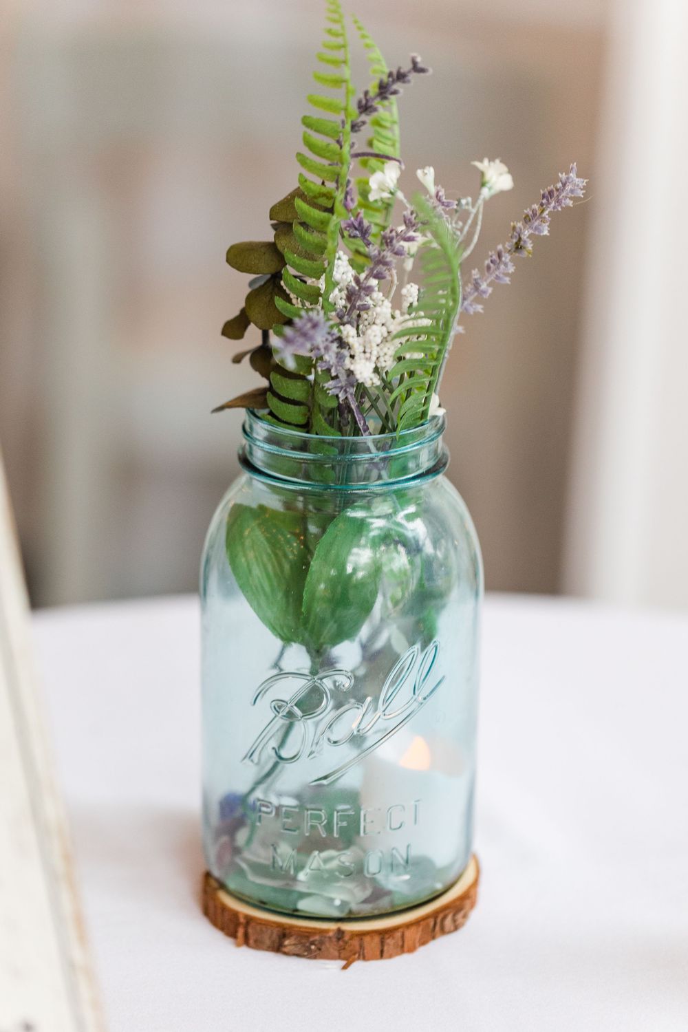 Blue mason jar centerpiece with ferns and white flowers on a rustic wooden coaster.