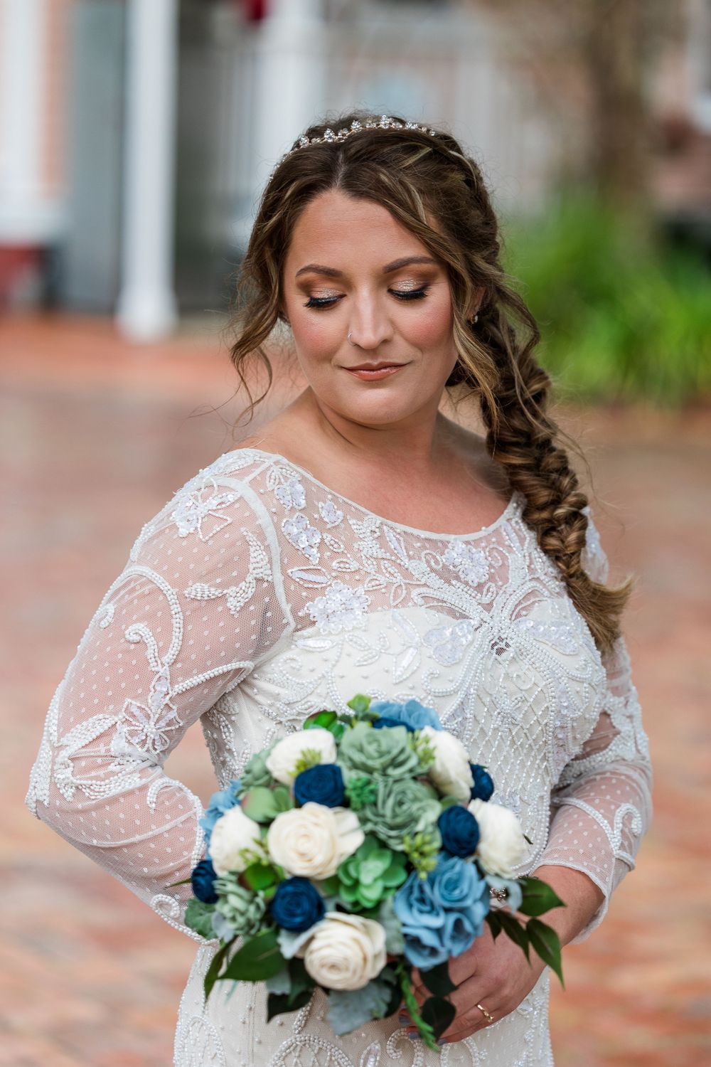 A bride in a lace long-sleeve wedding dress holds a beautiful bouquet of blue, white and green flowers.
