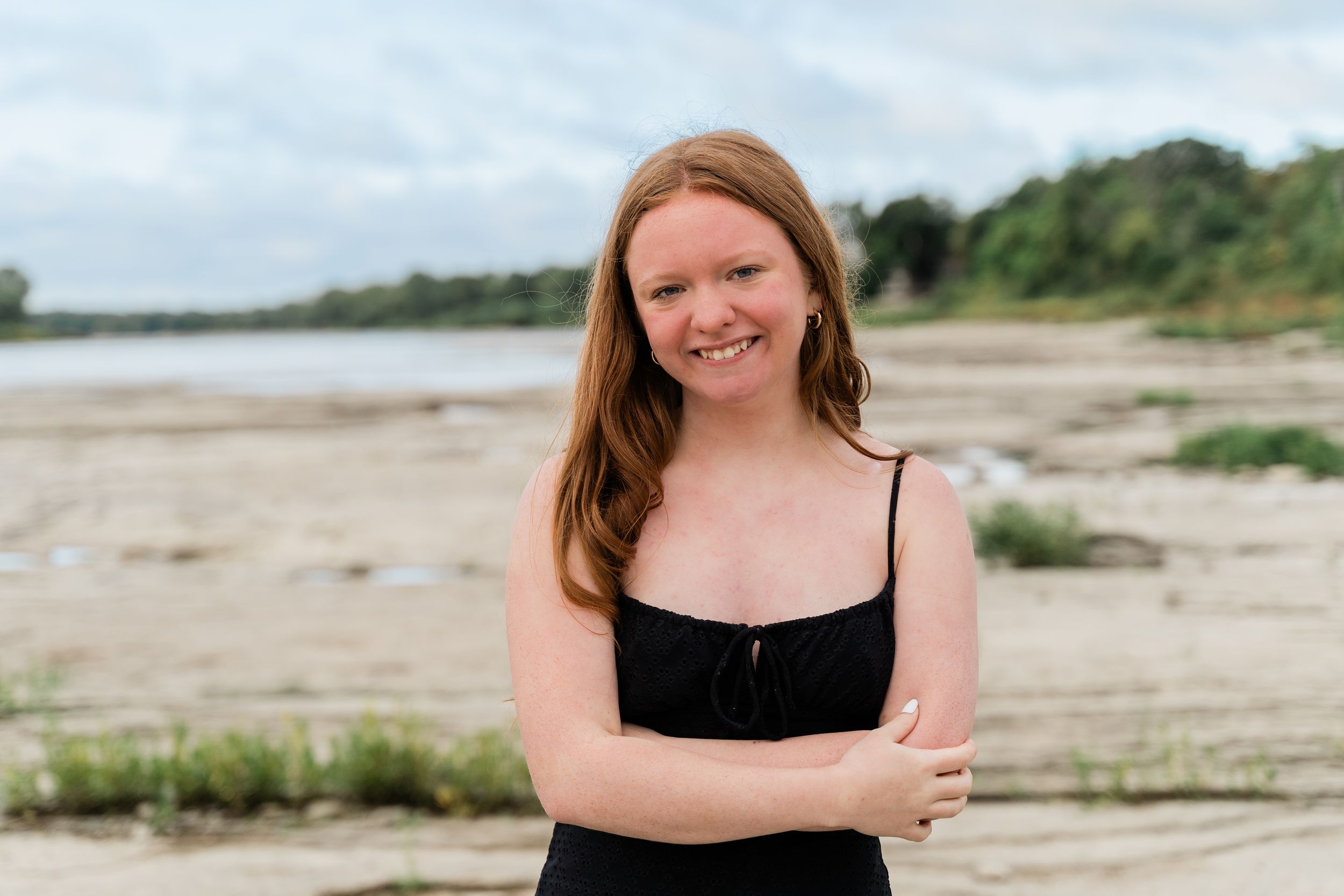 A woman in a black dress poses on a sandy beach with coastal vegetation and water in the background.