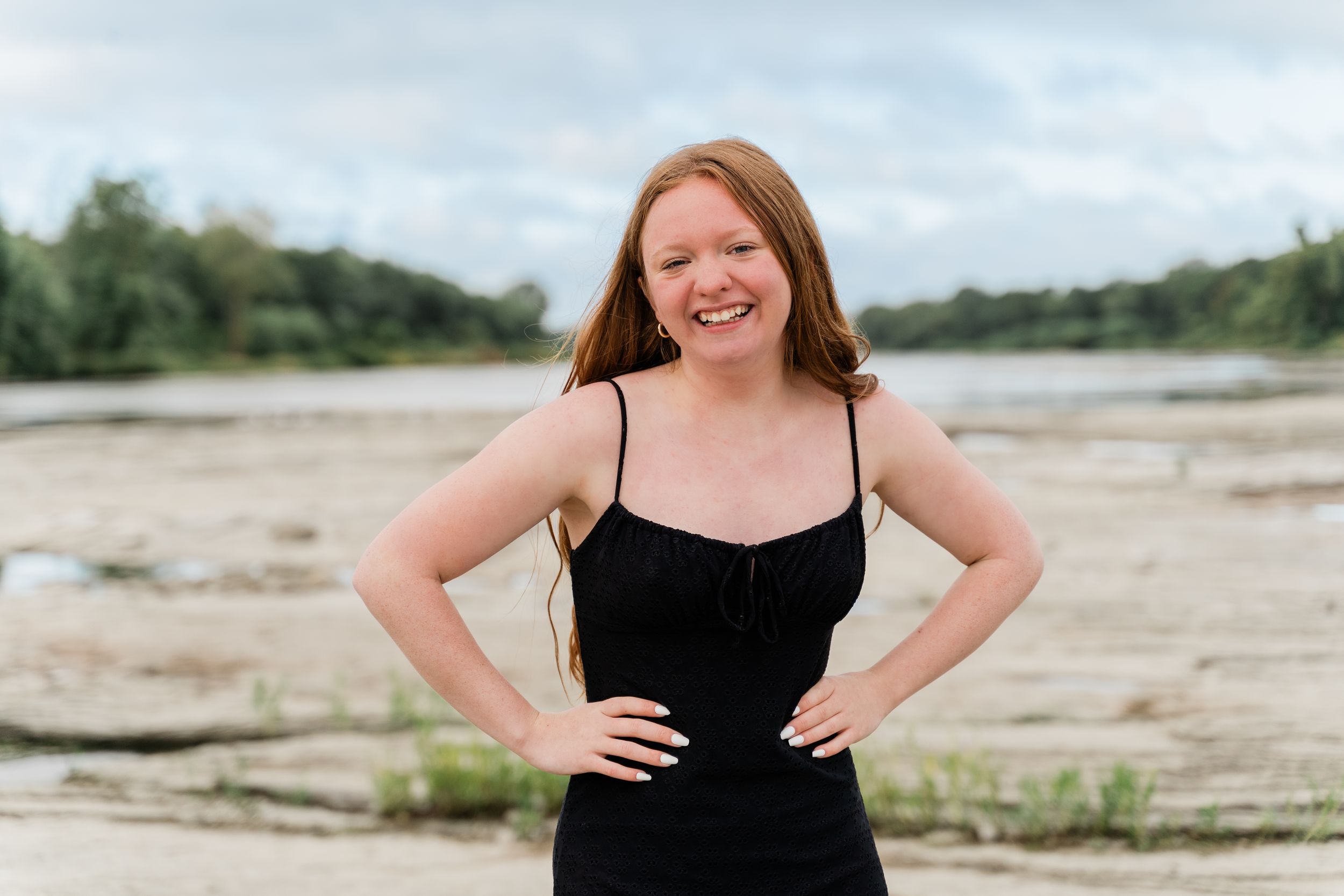 Woman in black dress poses confidently on sandy beach during summer photoshoot.