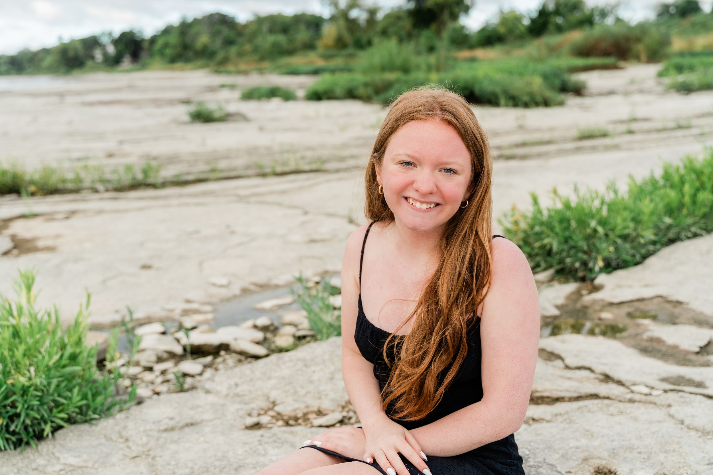 A person with long hair sits outdoors on rocky terrain surrounded by greenery on a sunny day.