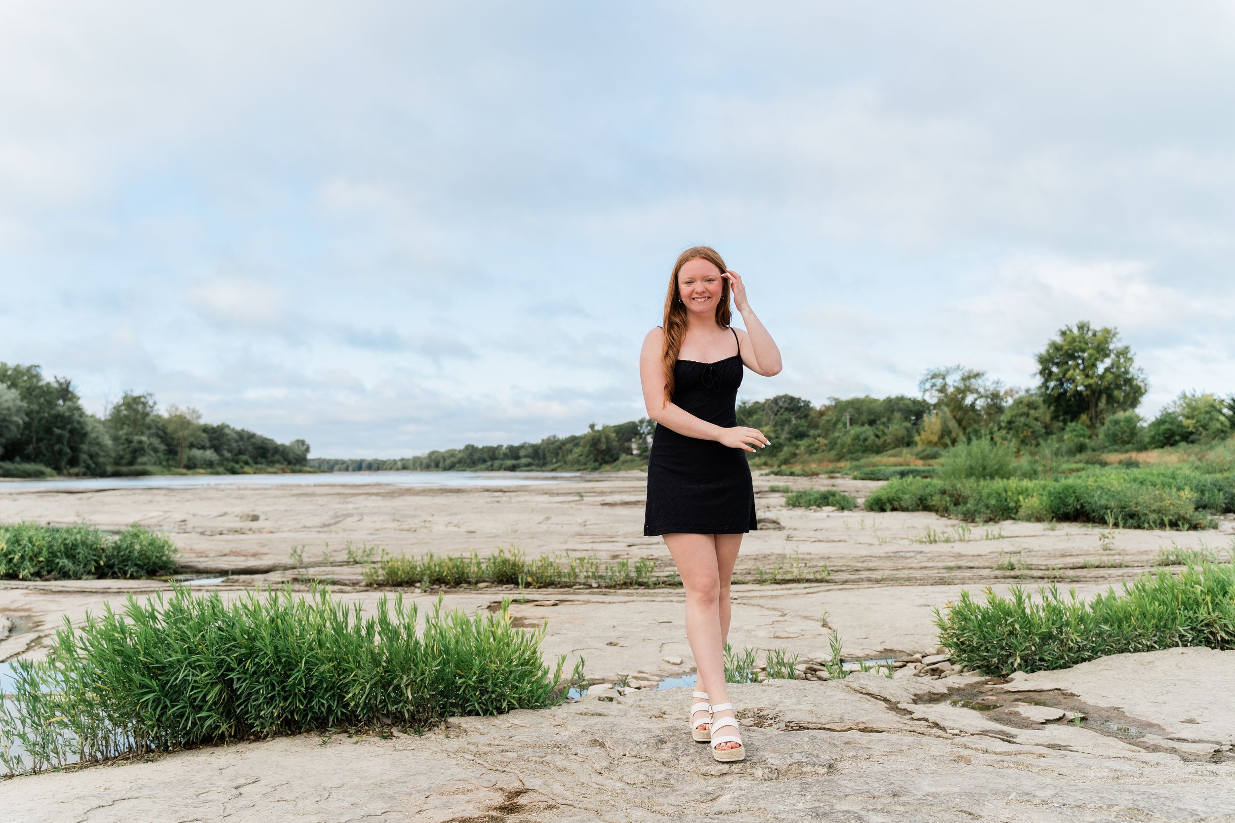 A dancer in a black dress performs graceful poses on a rocky beach landscape at sunset.