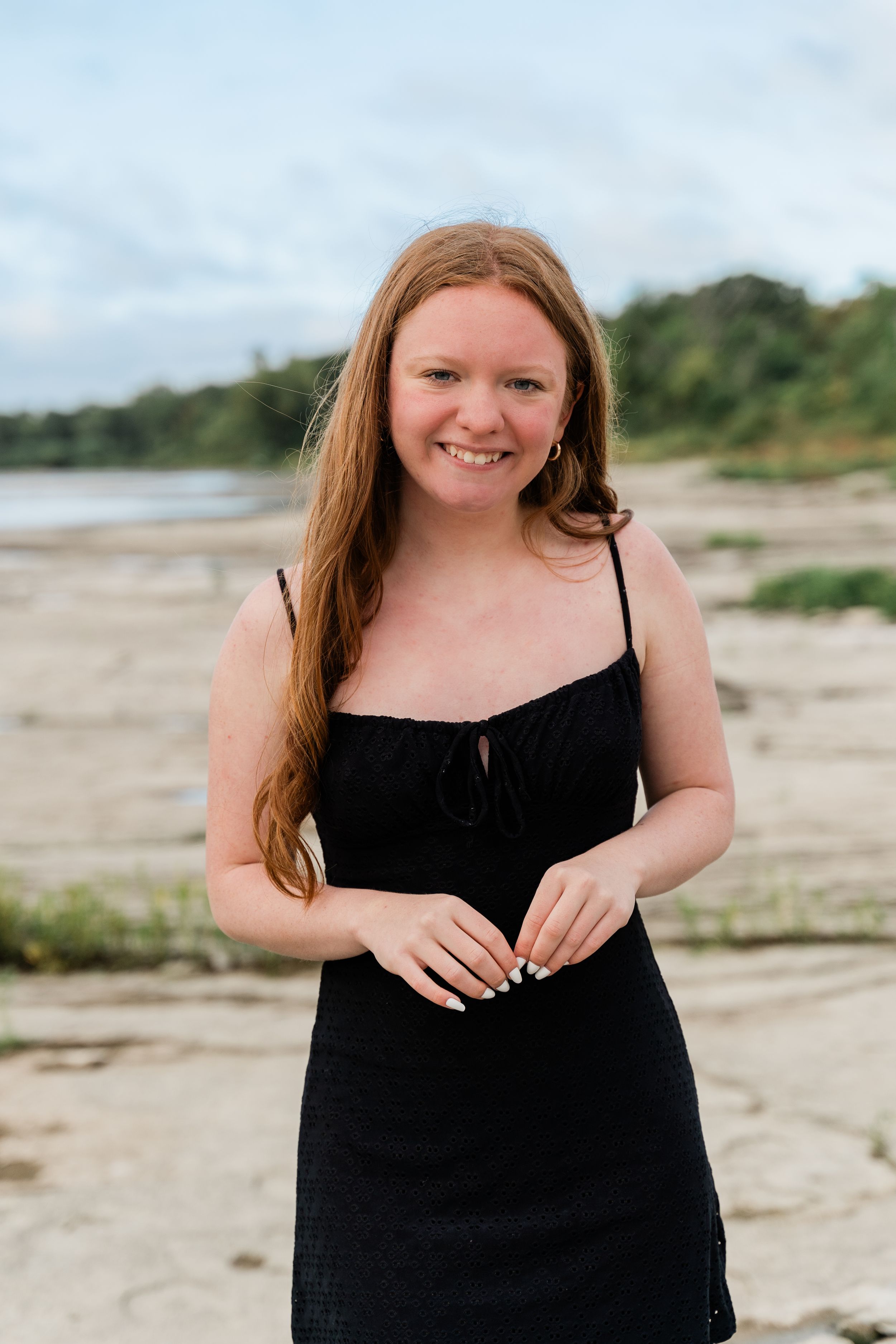 Person with long red hair wearing a black dress poses on a rocky beach with trees in the background.