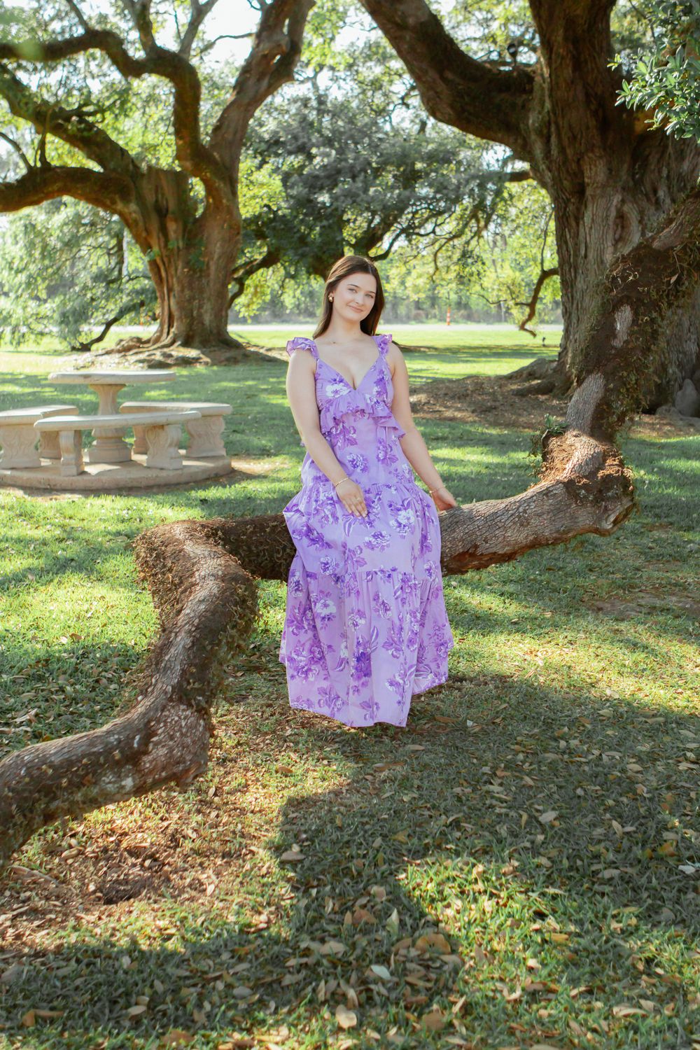 Young woman in purple floral maxi dress posing beside a large oak tree in a lush green park.
