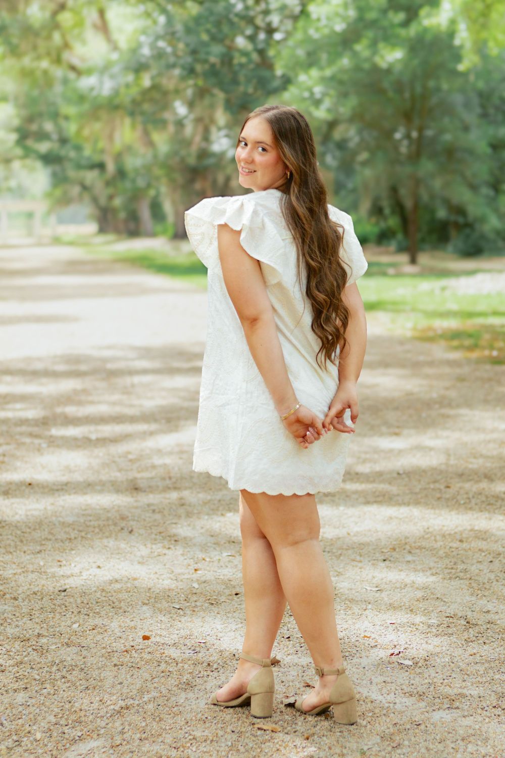 Young woman in white eyelet dress and tan heels standing on gravel path surrounded by green trees.