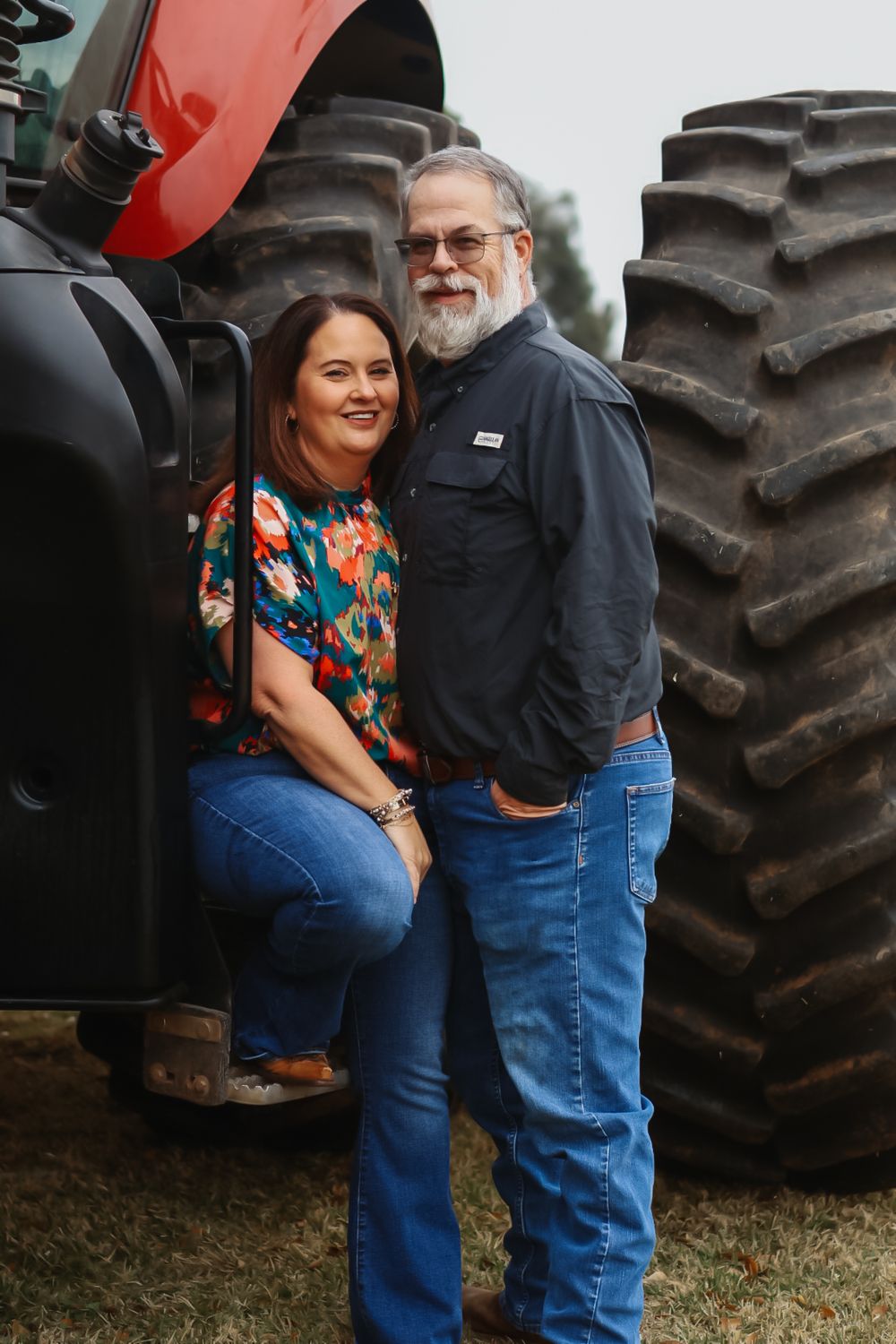 Couple embraces in front of large tractor tires in a romantic outdoor photoshoot.