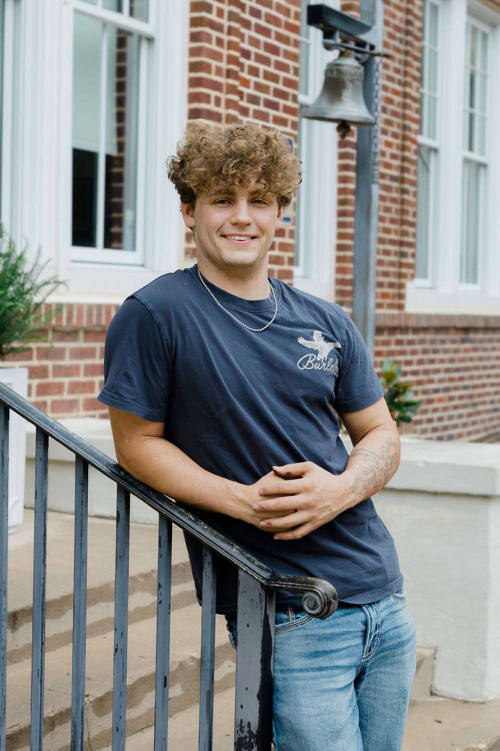 Young man with curly hair smiling in navy t-shirt and jeans, leaning on railing outside brick building.