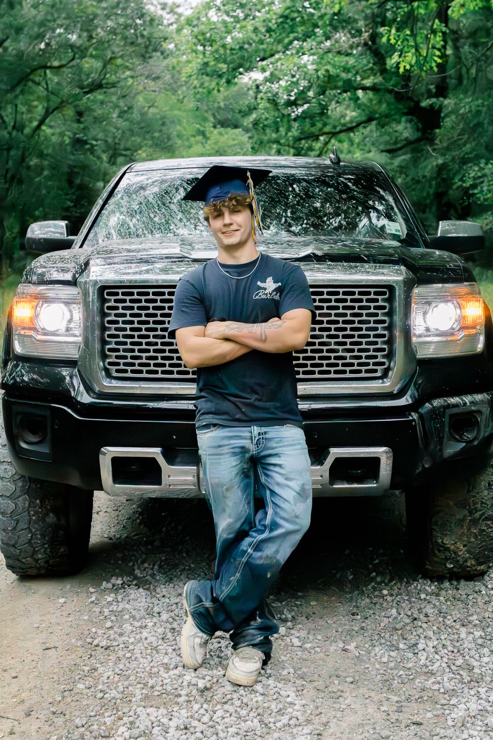 Young male graduate in cap and jeans poses arms crossed in front of black GMC pickup truck on gravel path.