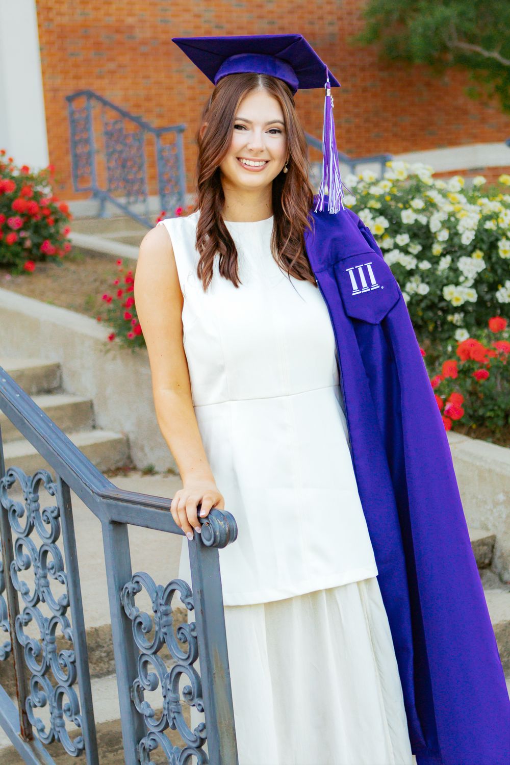 Smiling graduate in white dress holding purple cap and gown on outdoor staircase with flowers in background.