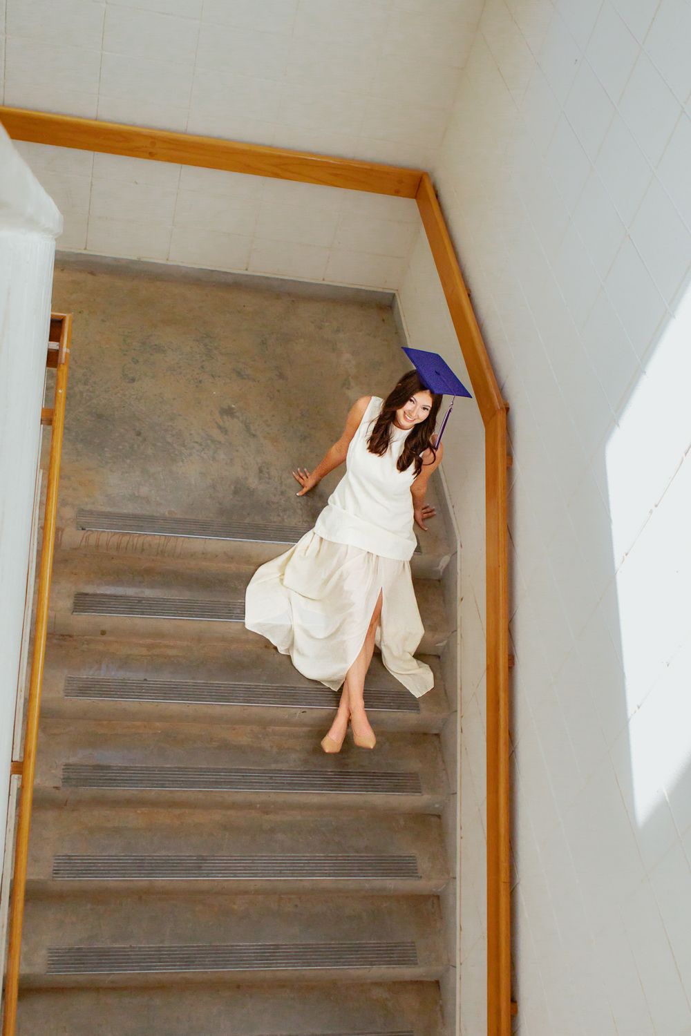 Woman in white dress holding blue umbrella descending indoor stairs, looking up at camera.
