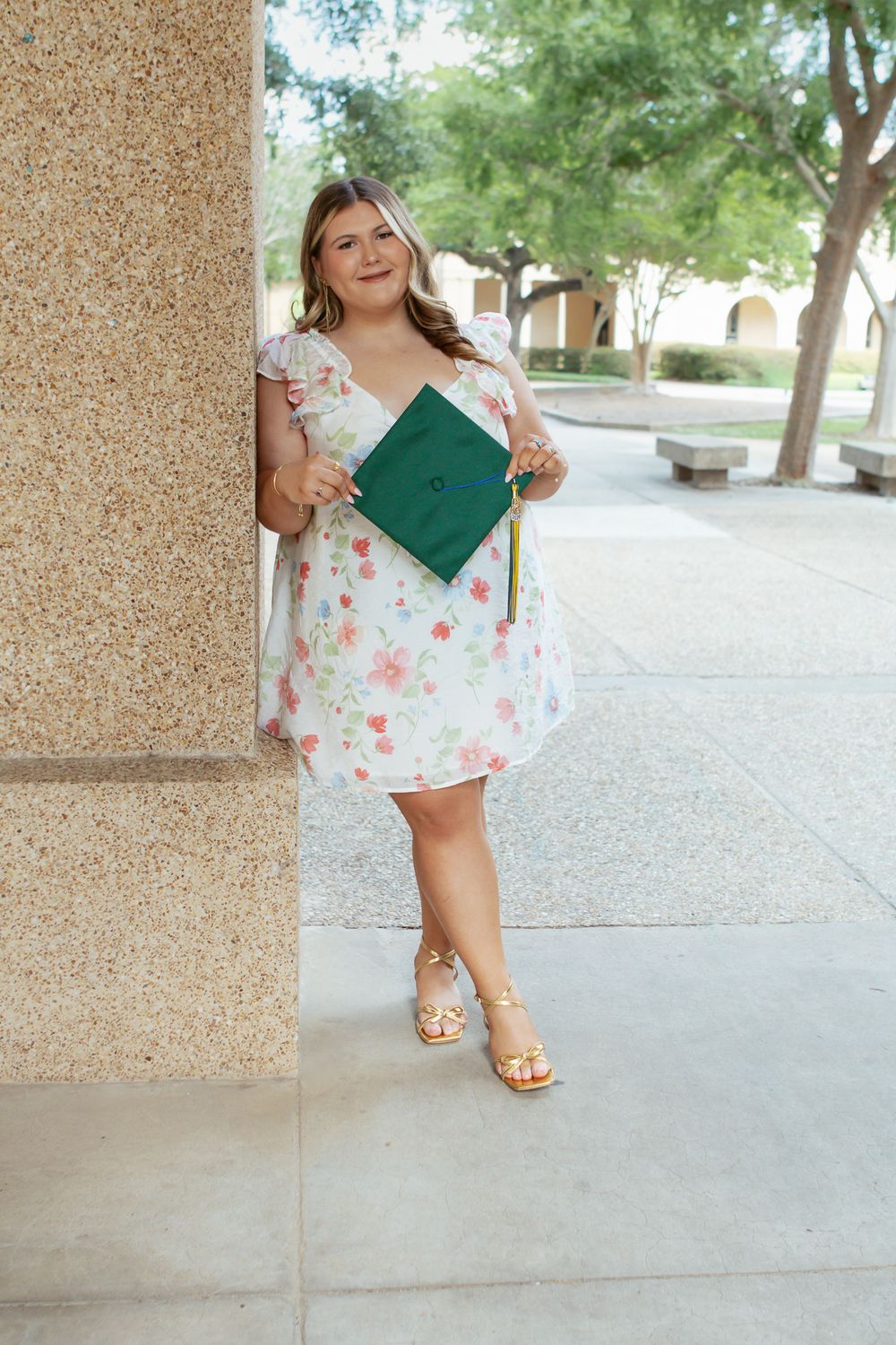 Young woman in floral dress holding green graduation cap, leaning against stone column on college campus.