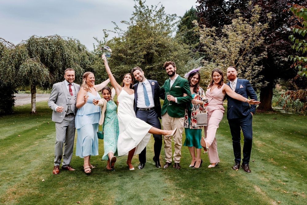 A lively wedding party poses together on a green lawn with trees in the background, showing joyful expressions and poses.
