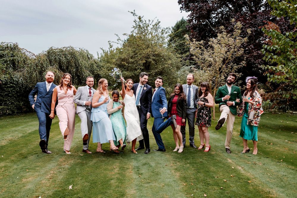 Wedding party lined up outdoors in a garden setting with trees and greenery in the background.