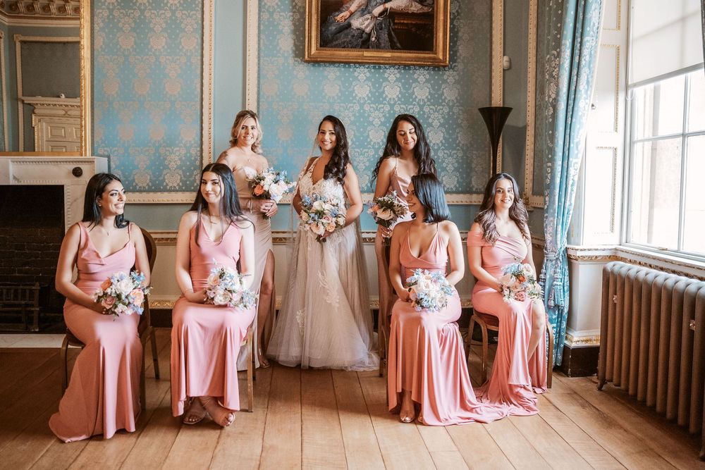 Bridesmaids in coral pink dresses gather in an elegant blue-walled room with large windows and ornate decor.