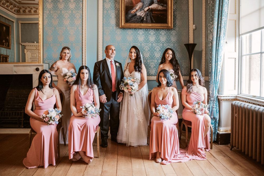 Bridesmaids in pink dresses gather in elegant blue wallpapered room with ornate decor.