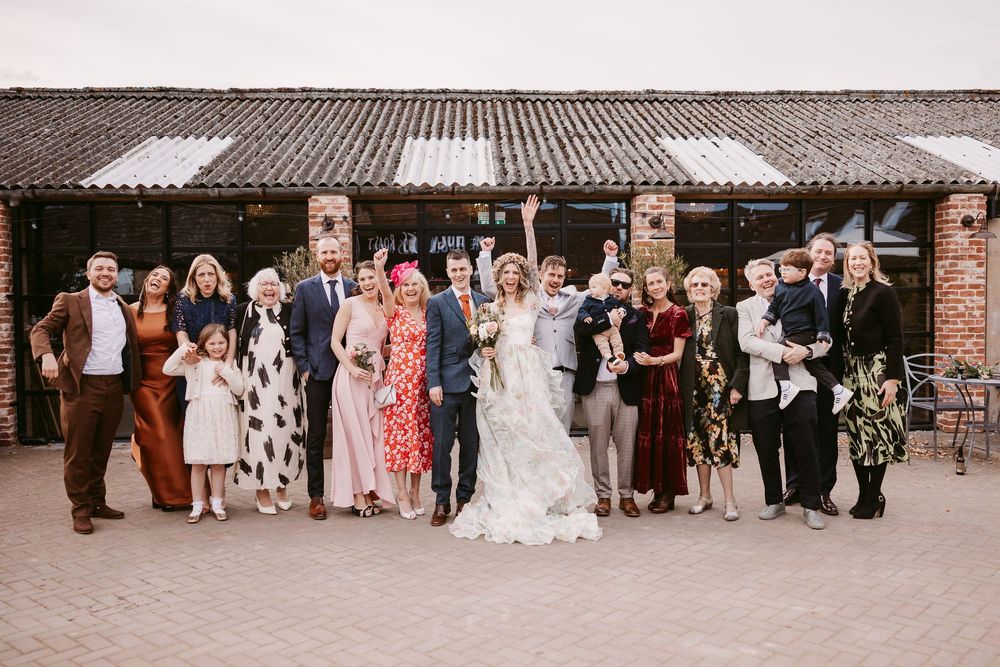 Wedding group photo lineup against a brick building backdrop with formal attire and mixed color dresses.
