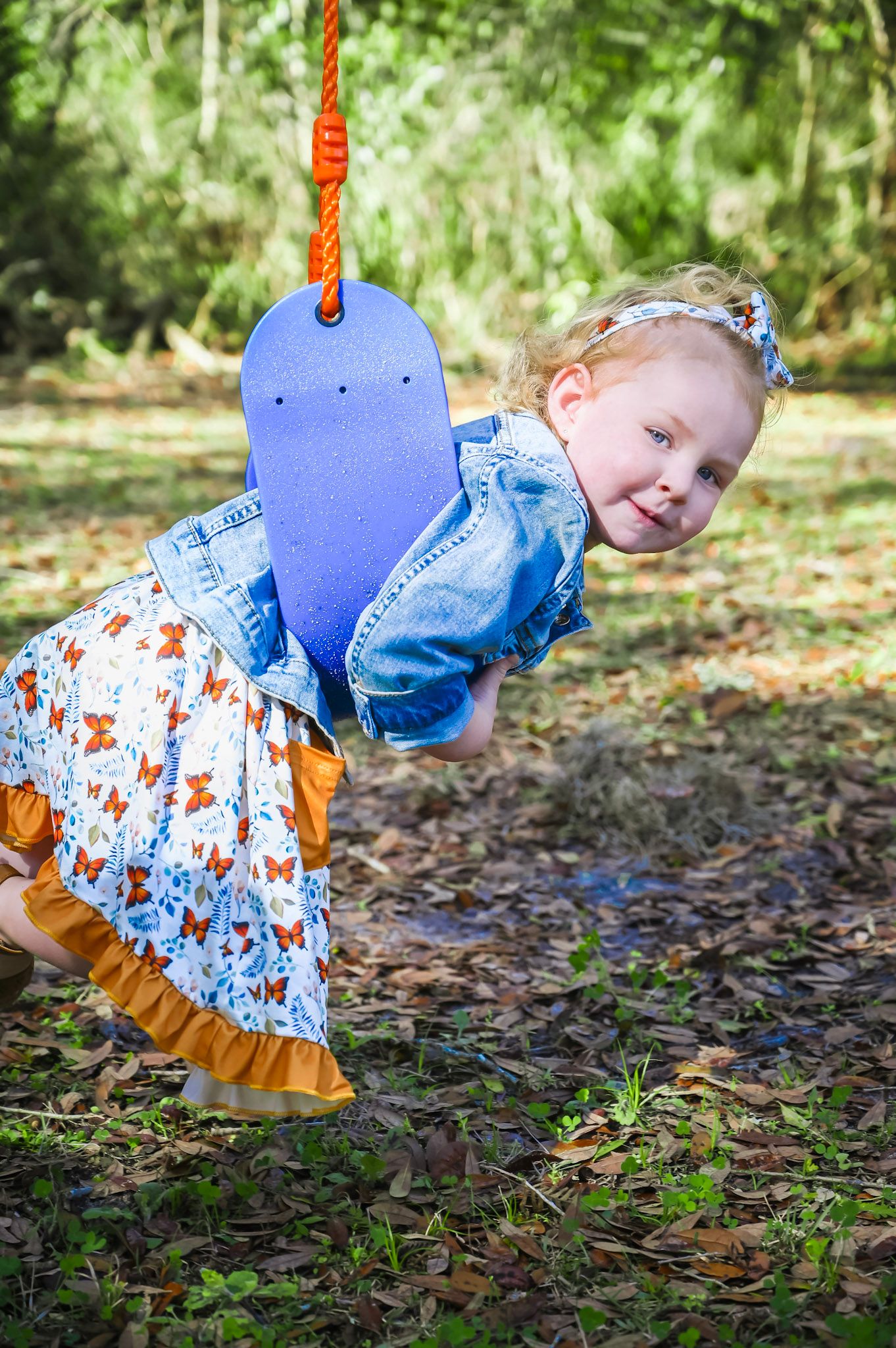 A child in a blue and white dress with orange accents plays in a forest setting with fallen leaves.