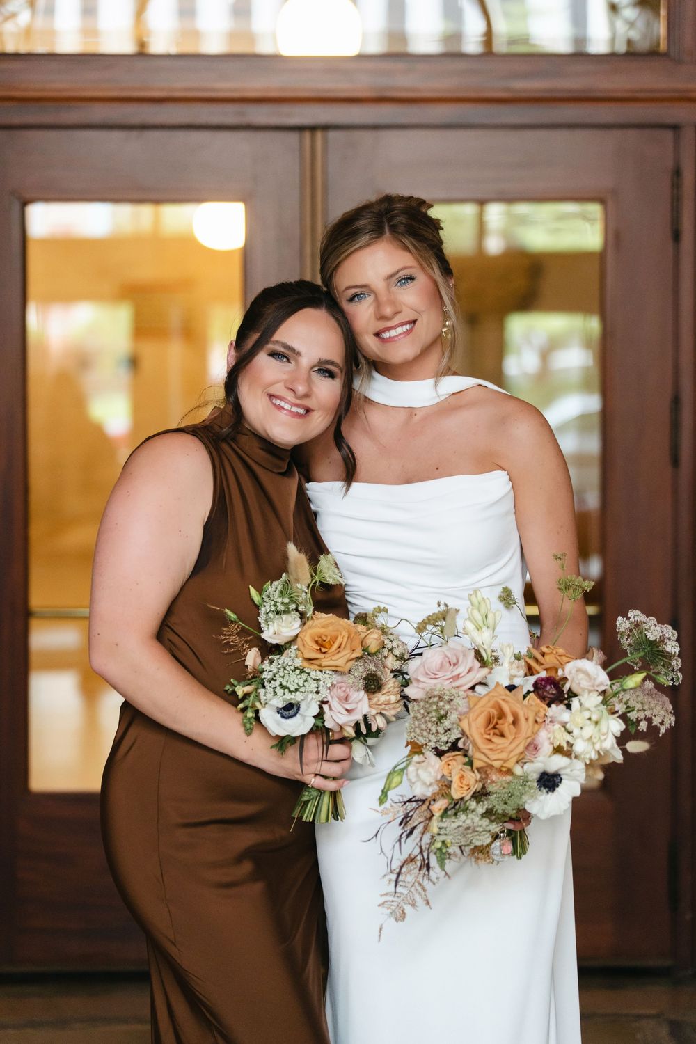 Bridesmaids in brown dresses hold peach and white floral bouquets next to bride in white gown.