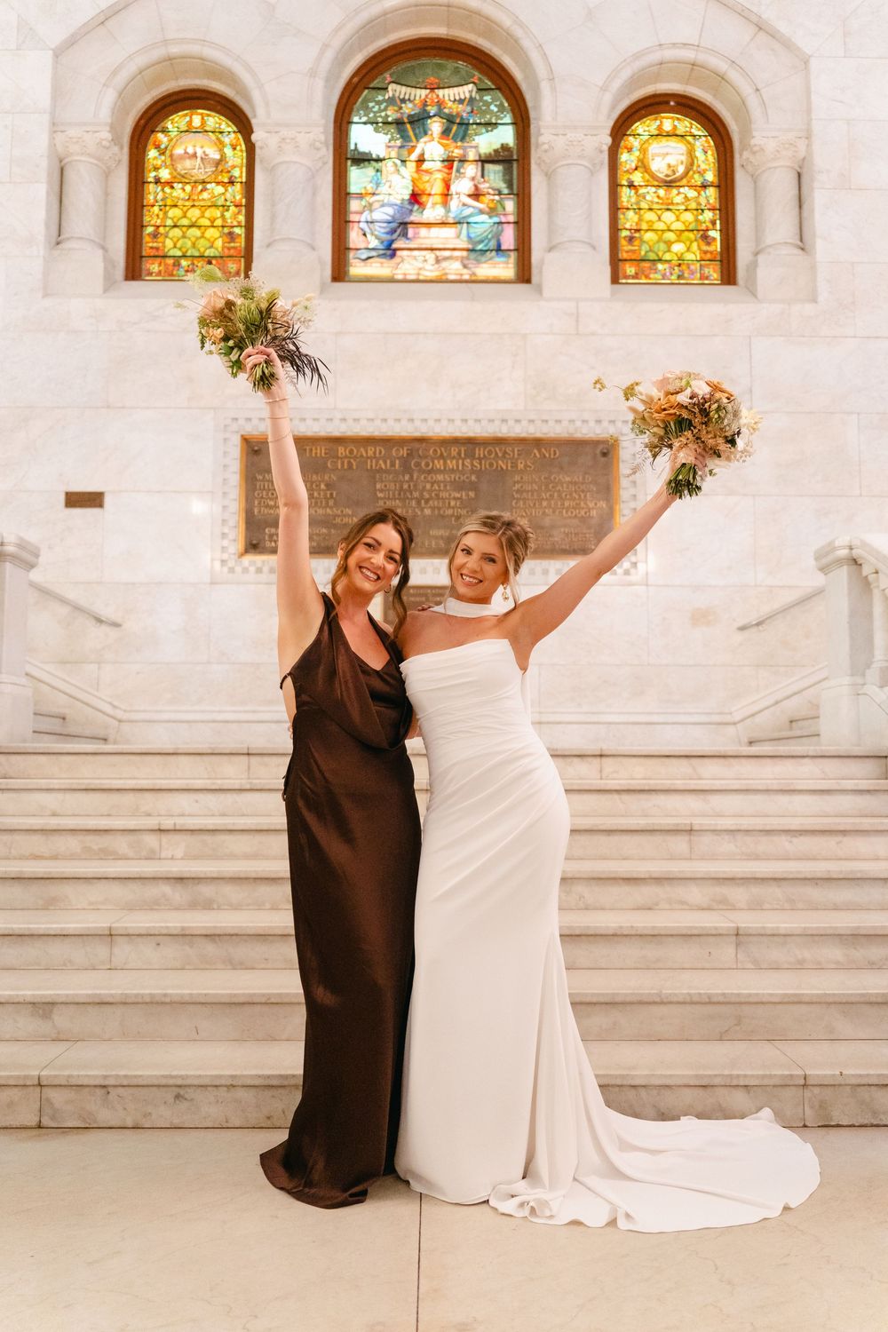 Two people in brown and white dresses pose playfully on marble steps in front of stained glass windows.