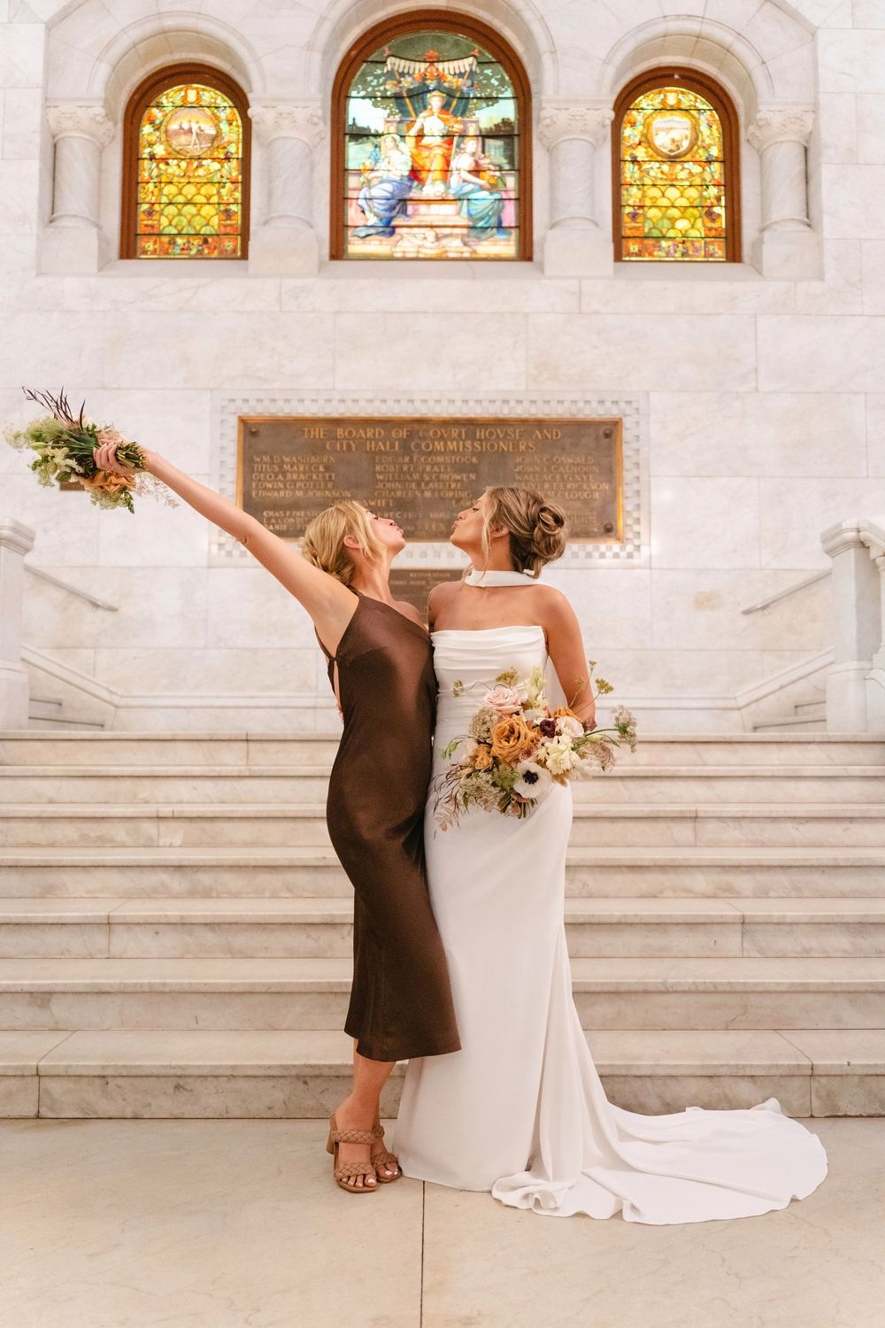 Wedding party poses dramatically on marble stairs with stained glass windows in background.