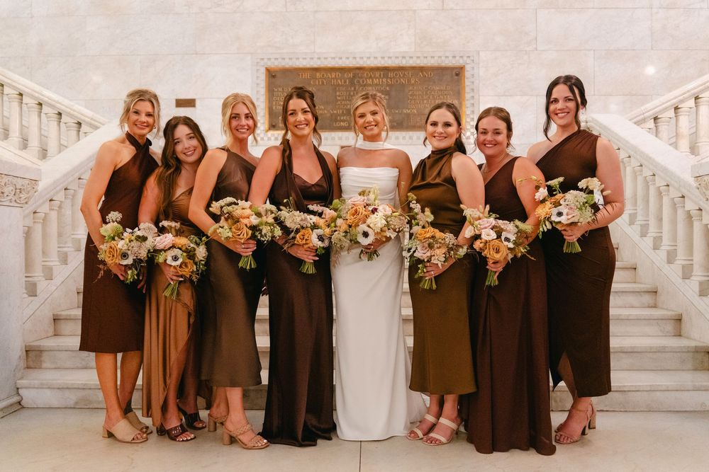 A group wearing brown bridesmaid dresses and holding floral bouquets pose together on white stairs.